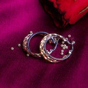 A pair of silver metallic dual-toned hoop earrings with a rhodium-based anti-tarnish coating, displayed on a pink fabric surface with a red rose petal nearby.
